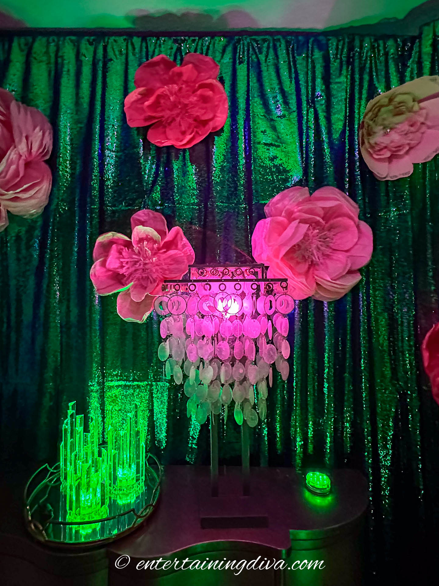 A decorative scene with large pink flowers, a crystal chandelier with pink light, green sequin drapes, and glowing green props.