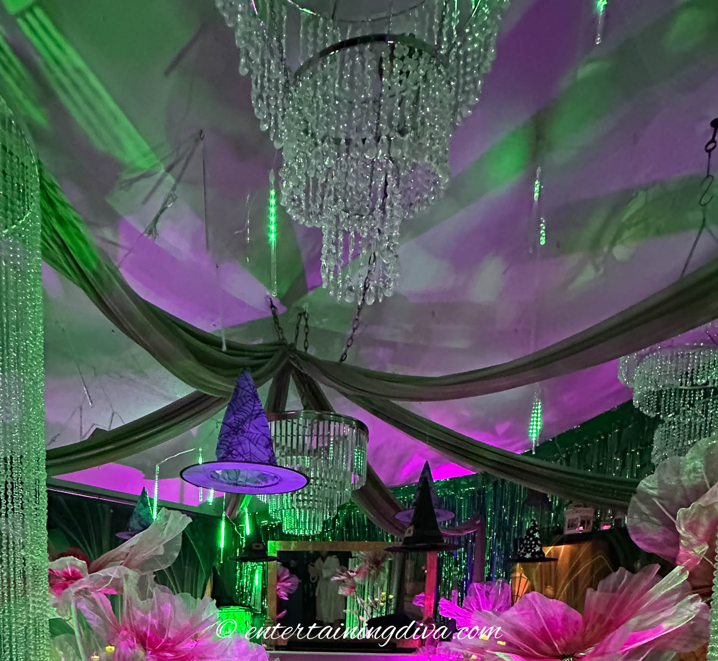 Decorated ceiling with crystal chandeliers, draped fabric, witch hats, and green and purple lighting, creating a festive atmosphere.