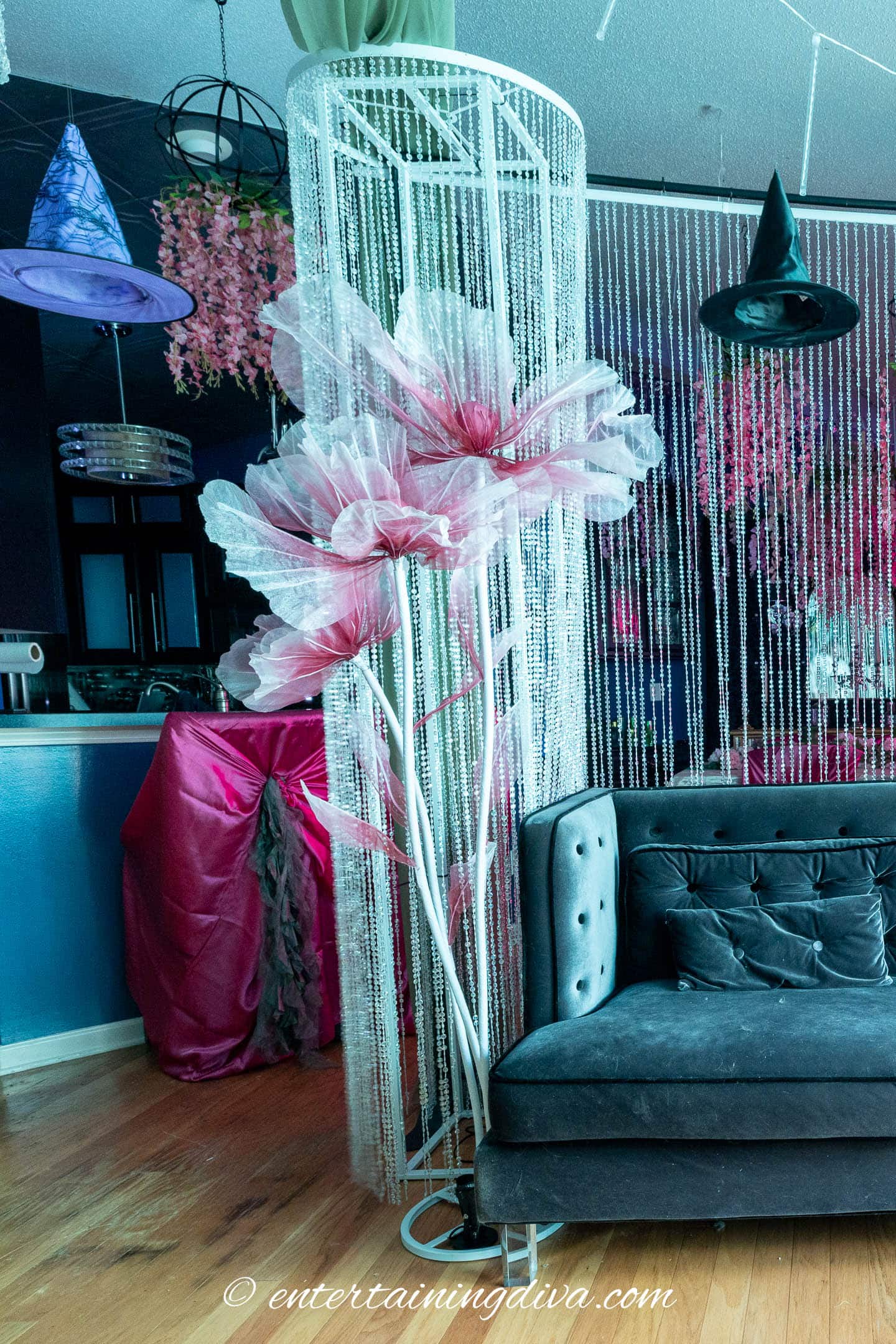A modern living room with a black couch, tall pink and white artificial flowers, beaded curtain, and witch hats hanging from the ceiling.