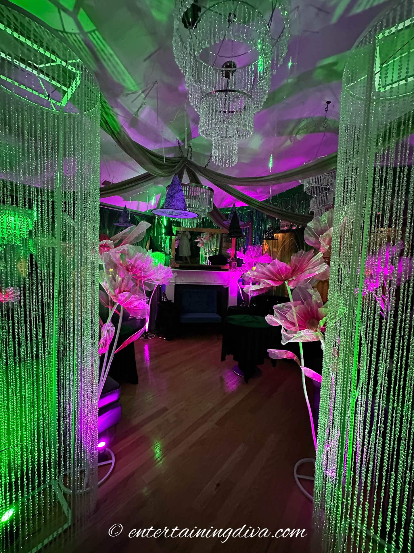 A room decorated for a party with green and purple lighting, hanging crystal bead curtains, oversized floral arrangements, and chandeliers.