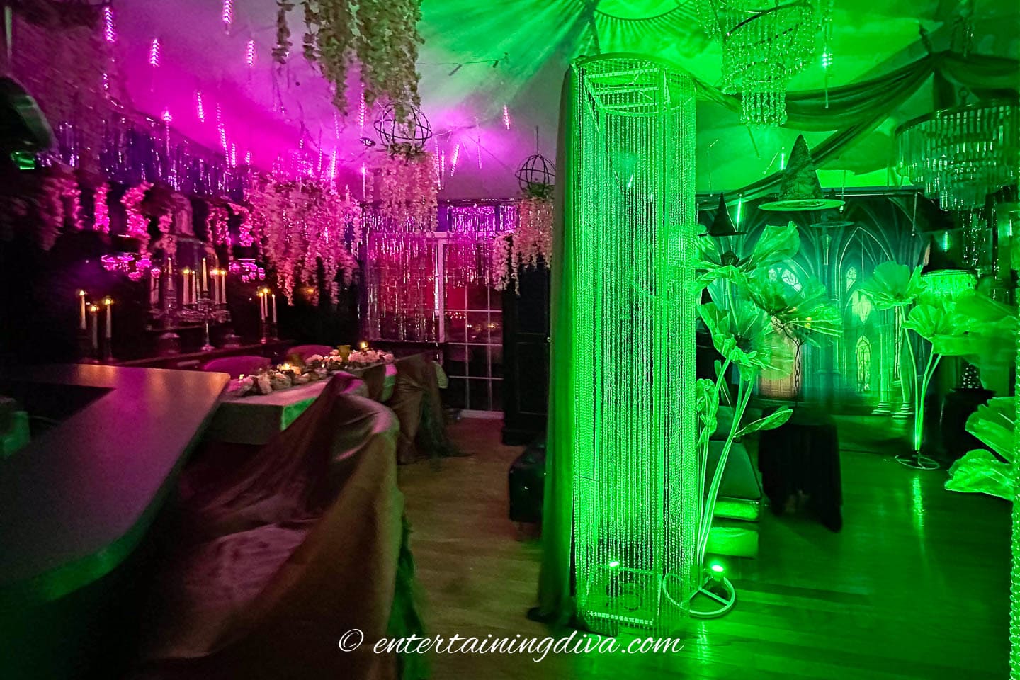 A room decorated for a party with green and purple lighting, hanging crystals, candles, and large artificial plants.