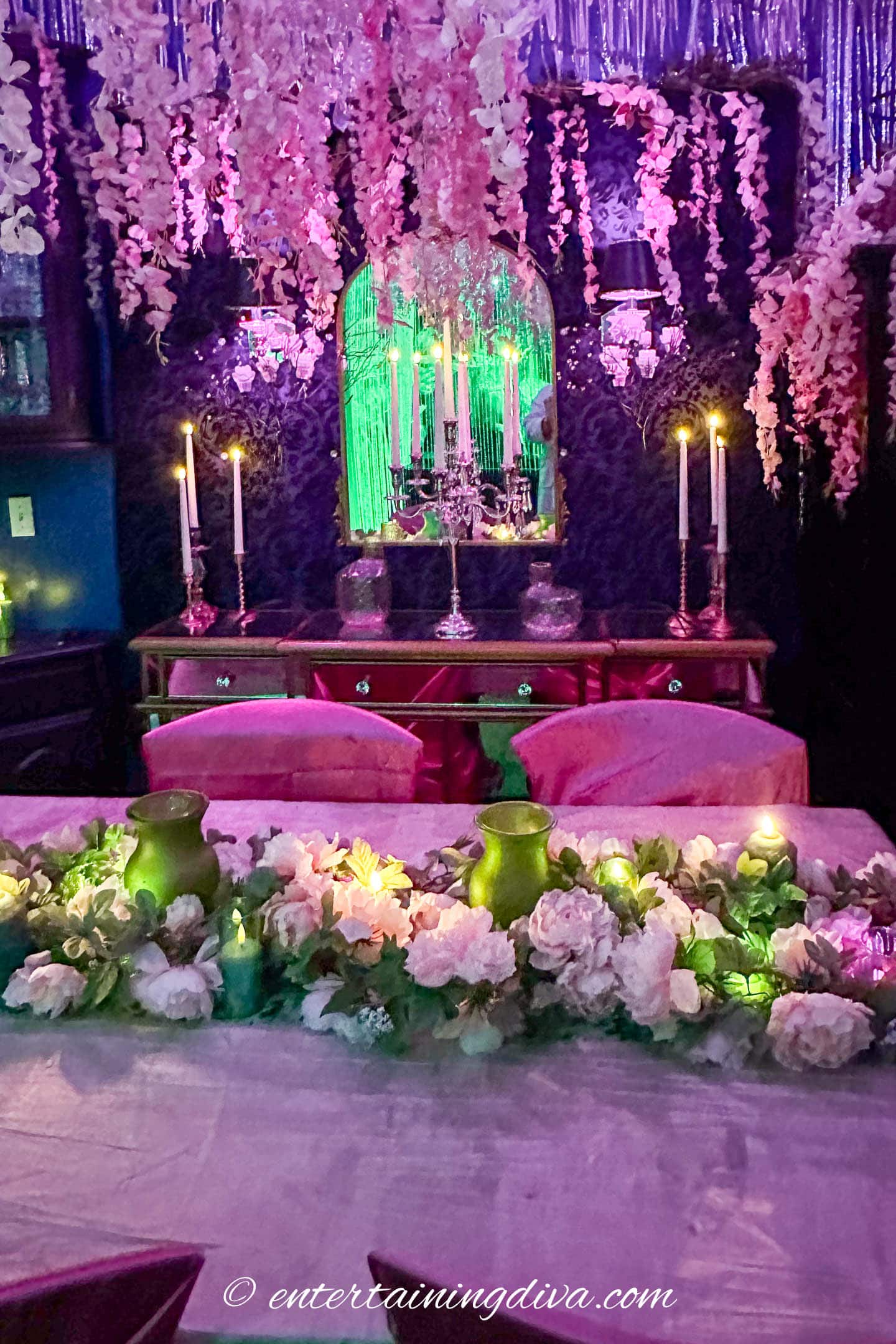 A dining table with a floral centerpiece, surrounded by candles and greenery, is set in a dimly lit room with purple and green lighting and hanging flowers.