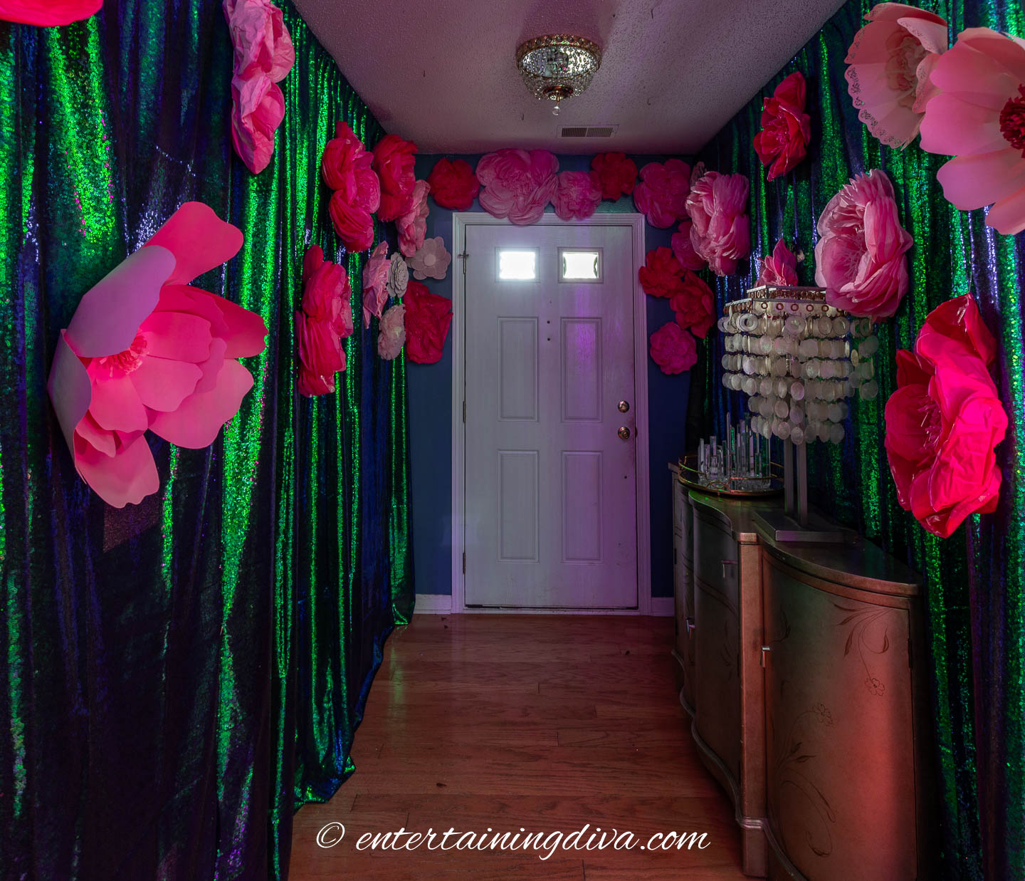 A hallway decorated with large pink paper flowers and shiny green curtains, leading to a white front door with a chandelier and a small mirrored table on the right.