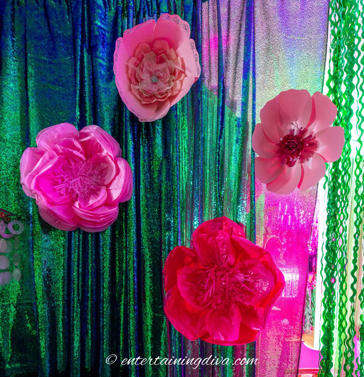 Four large pink paper flowers are displayed on a green and pink iridescent backdrop with a shimmery texture.