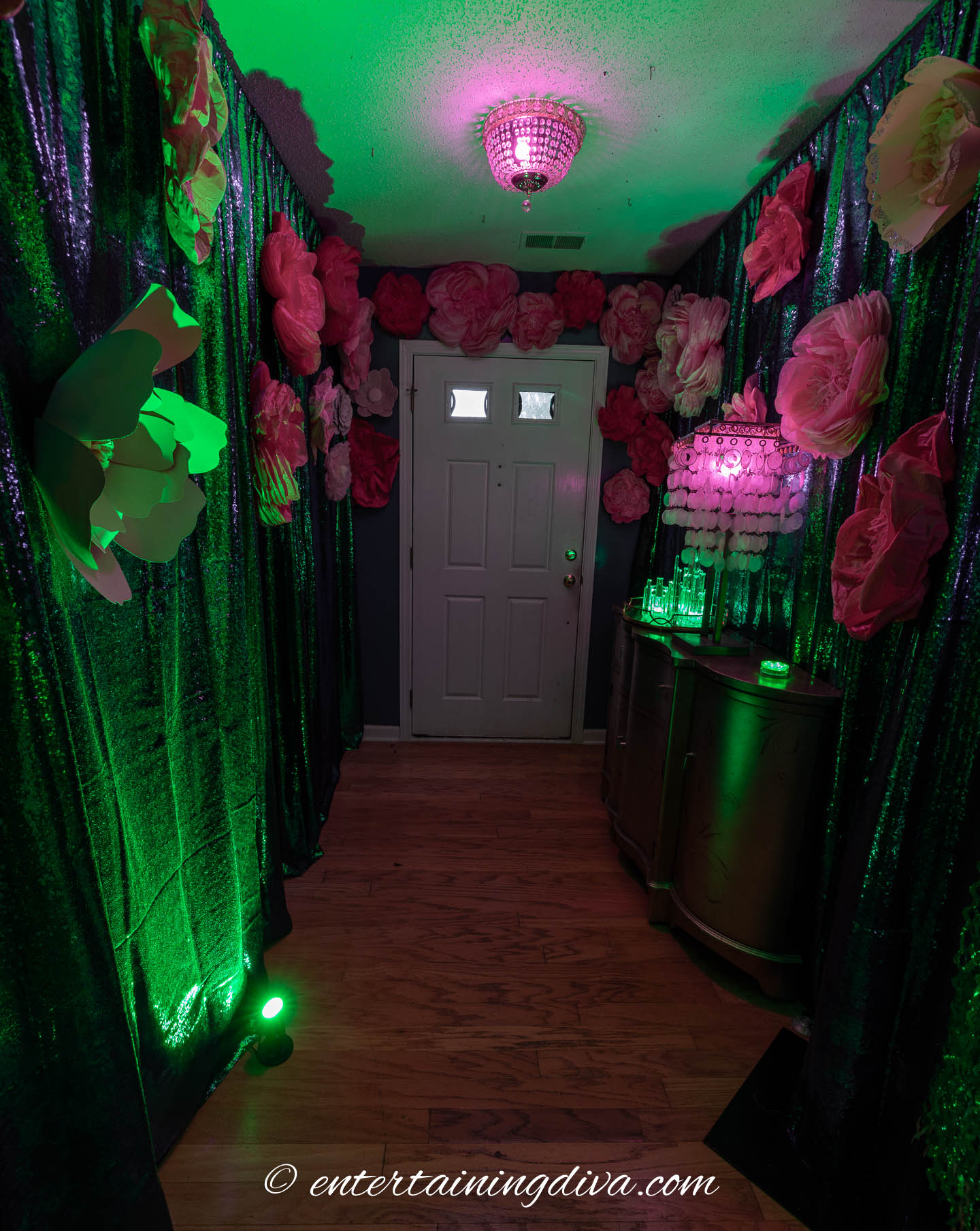 A hallway decorated with large paper flowers, green and pink lighting, and a small table with candles under a pink-lit ceiling light.