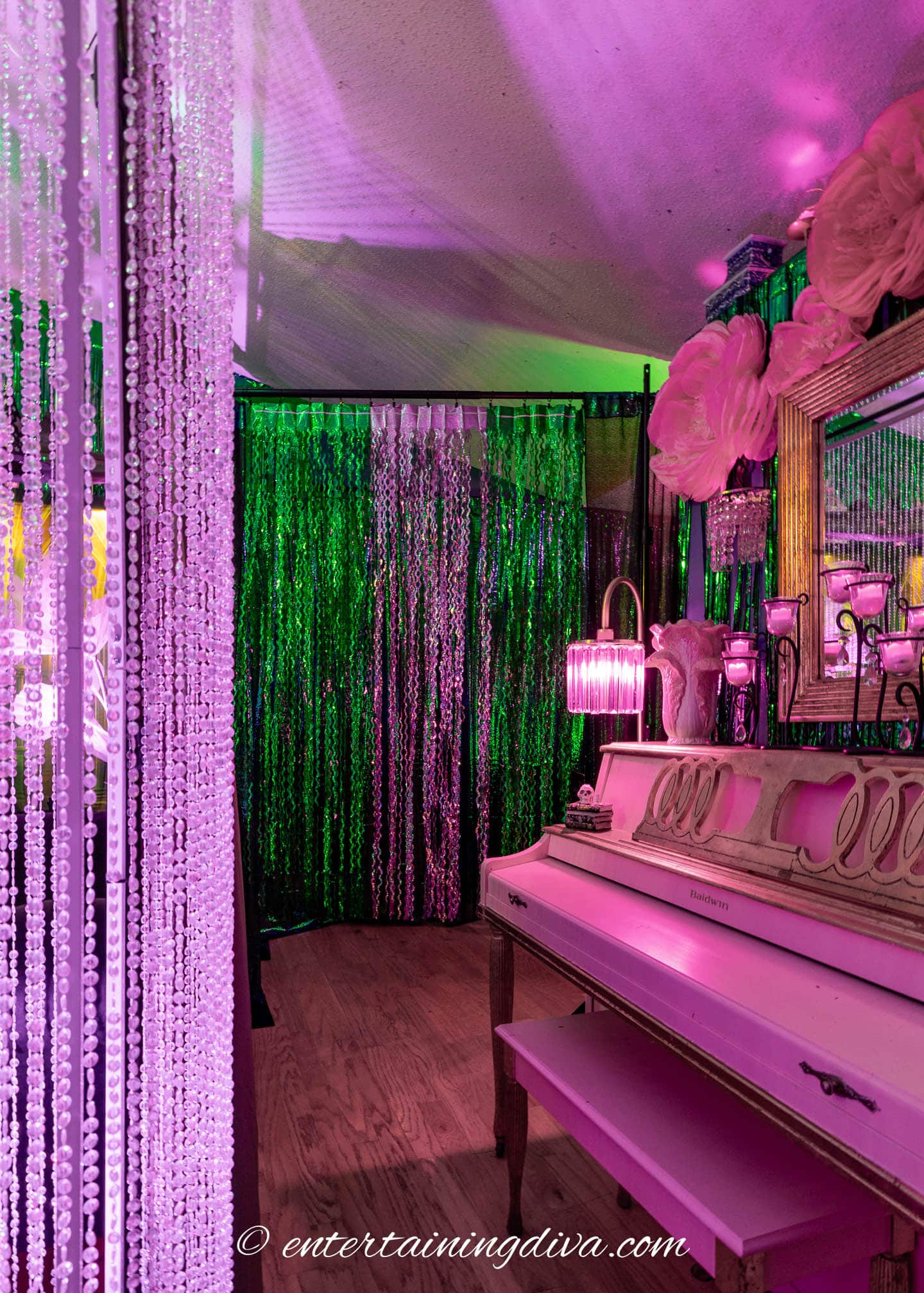 A white piano and bench sit beside a mirror and lamp, surrounded by colorful beaded curtains and vibrant pink and green lighting.