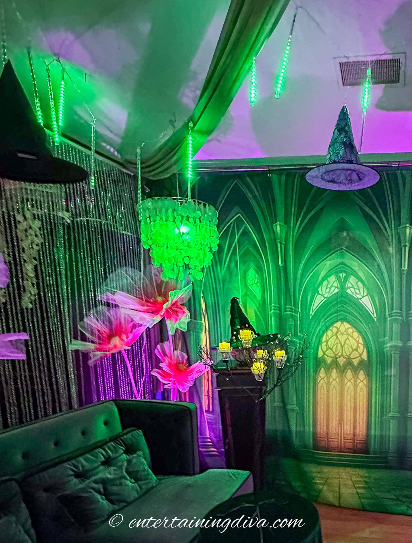 A room decorated with green lighting, hanging witch hats, a chandelier, large flowers, and a Gothic-style backdrop featuring arched windows.