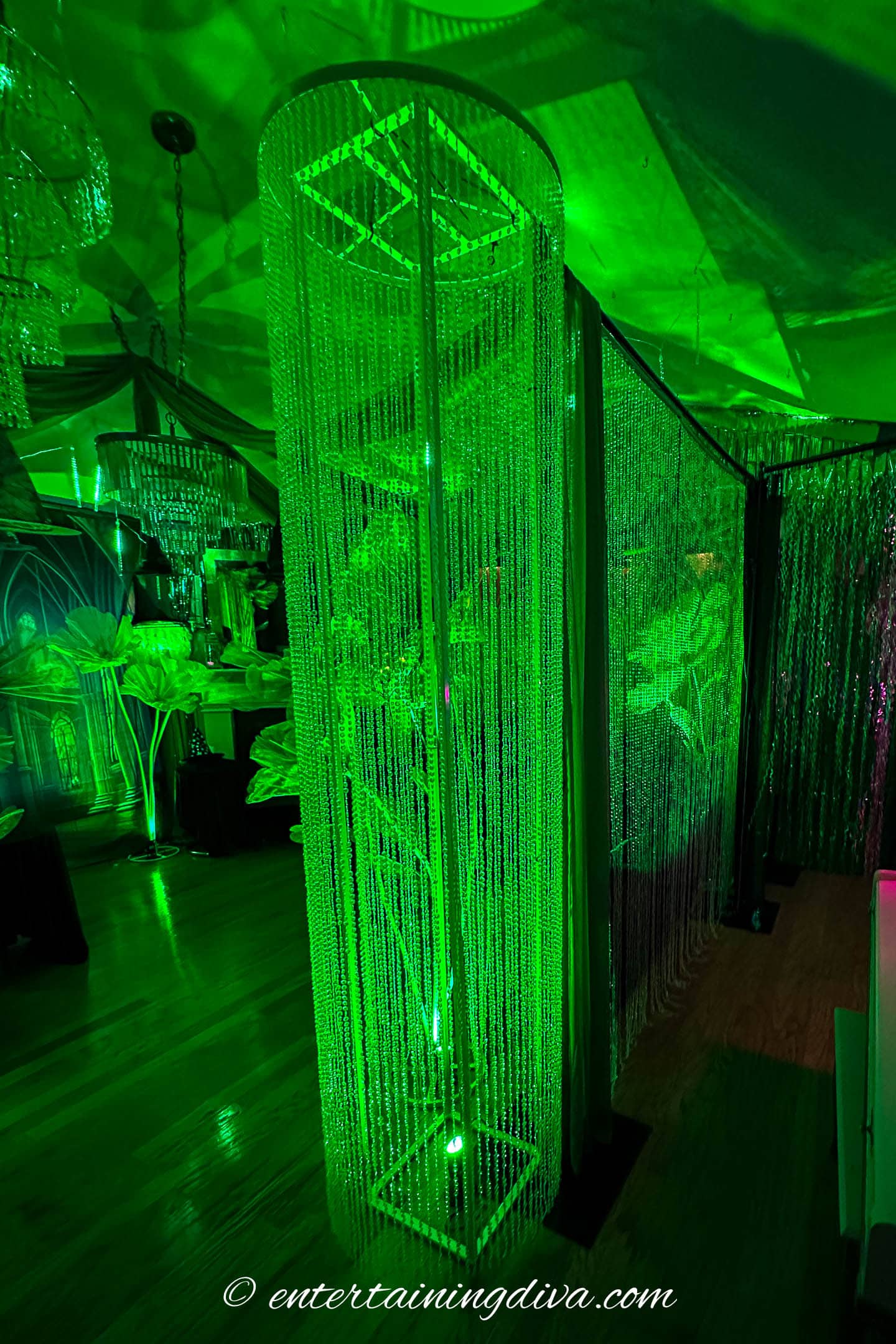 Tall rectangular column covered in hanging crystal strands, illuminated by green lighting in a decorated indoor space.