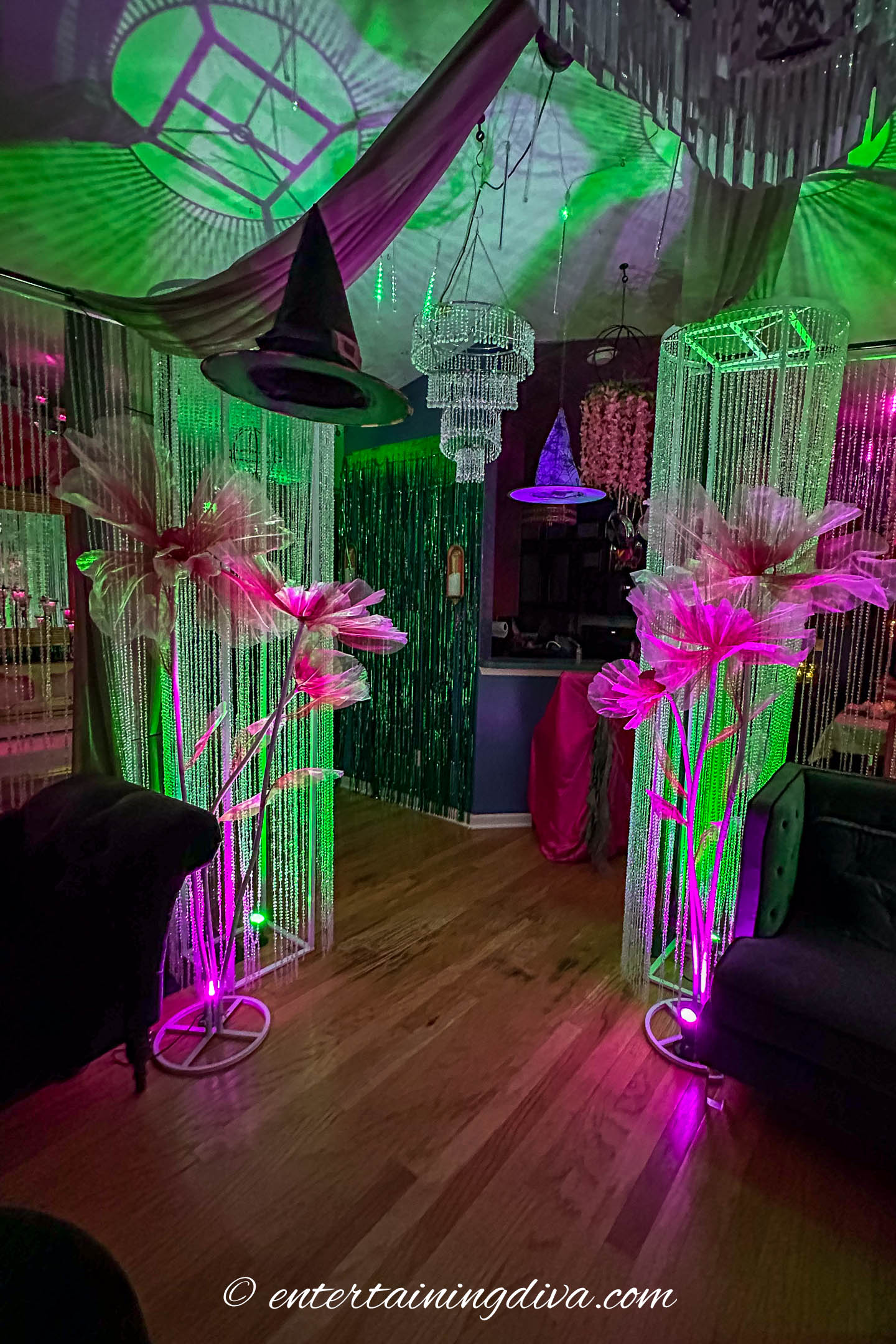 Room decorated with oversized pink flowers, beaded curtains, chandeliers, green and purple lighting, and a hanging witch hat for a whimsical party atmosphere.