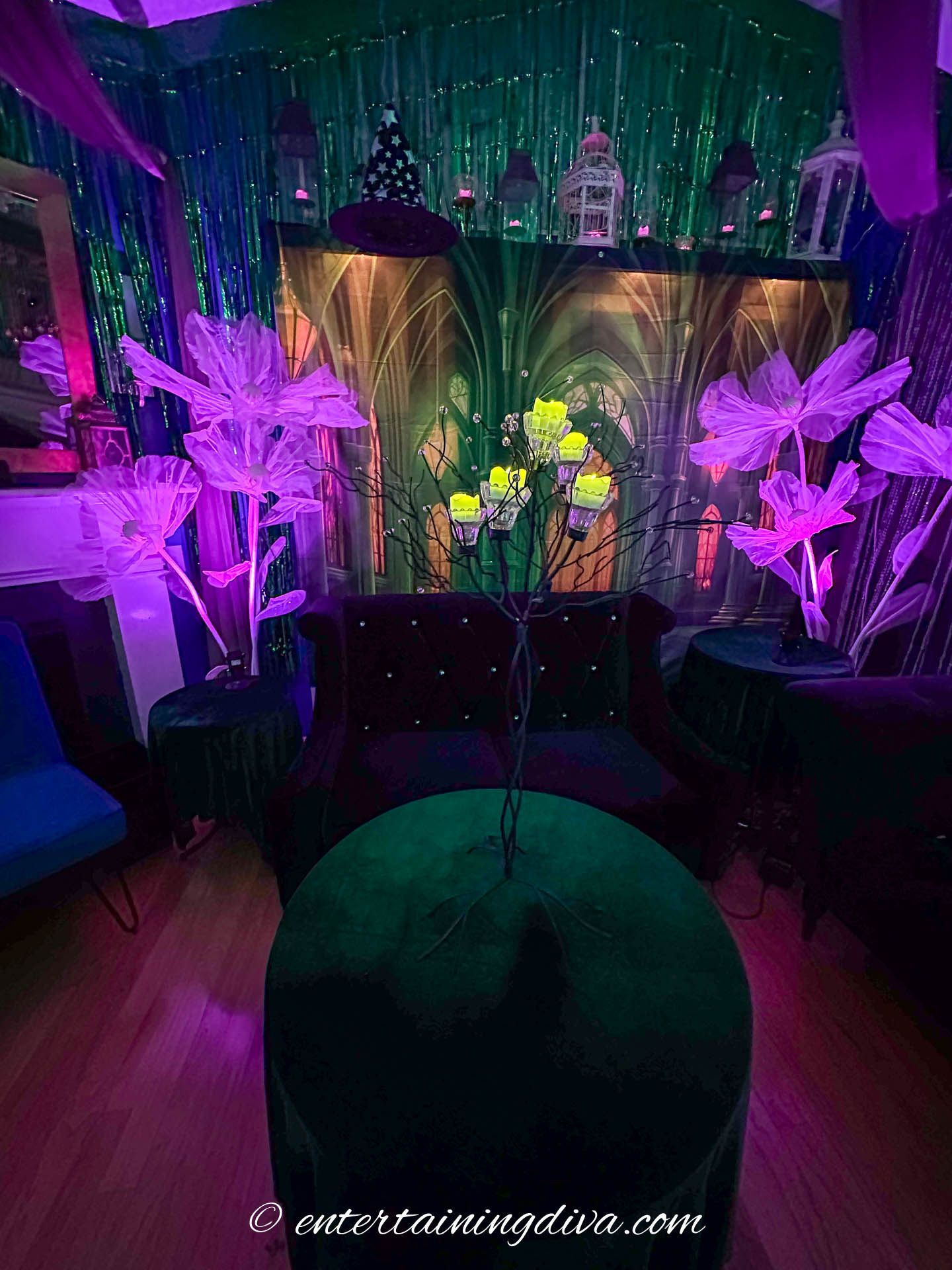 A room decorated with glowing, oversized flowers, a green round table, velvet chairs, and a backdrop of gothic arches, illuminated by colorful lights.