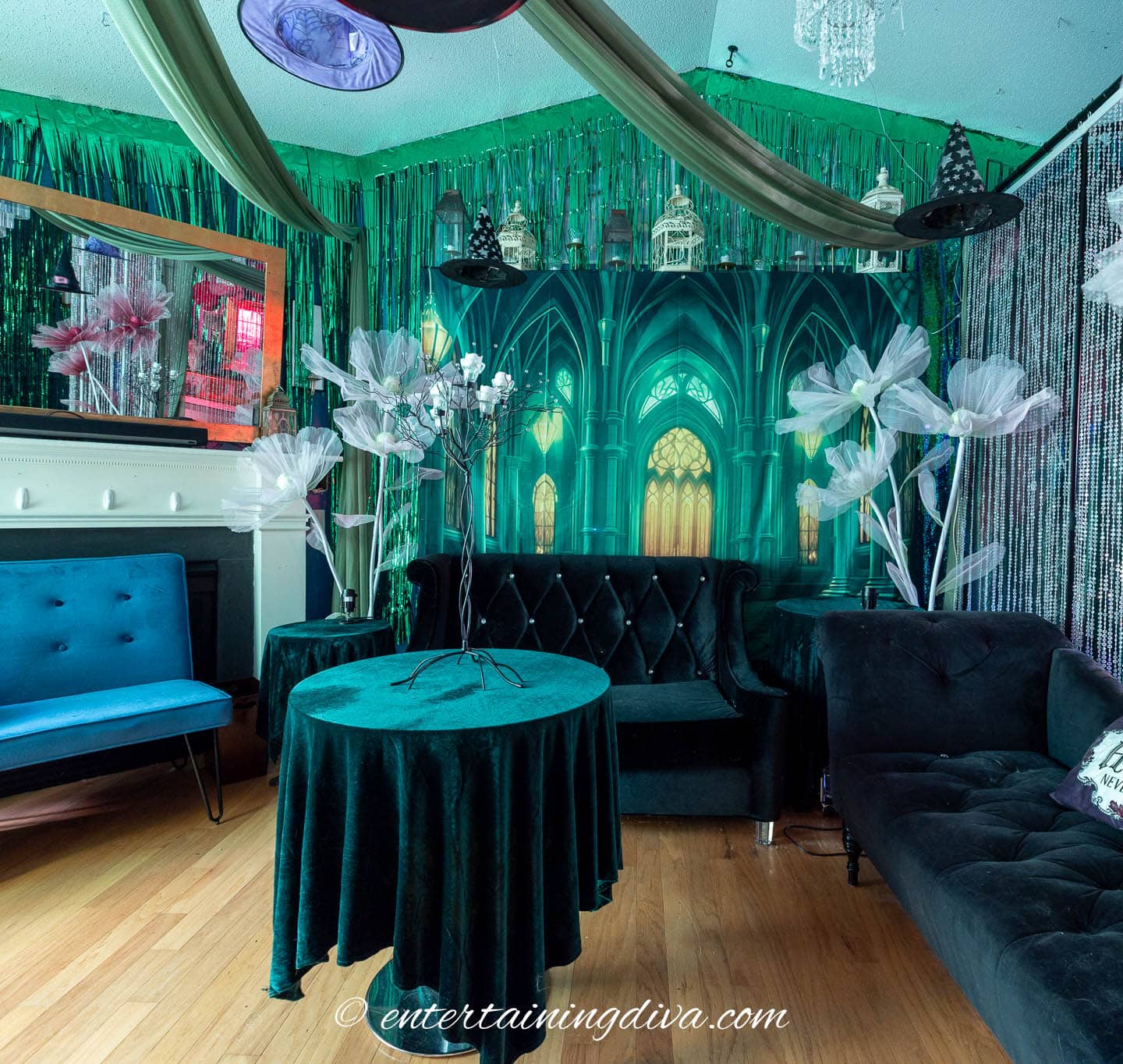Room decorated with green gothic arch backdrop, velvet furniture, faux white flowers, and metallic fringe, creating a whimsical, fantasy-inspired atmosphere.