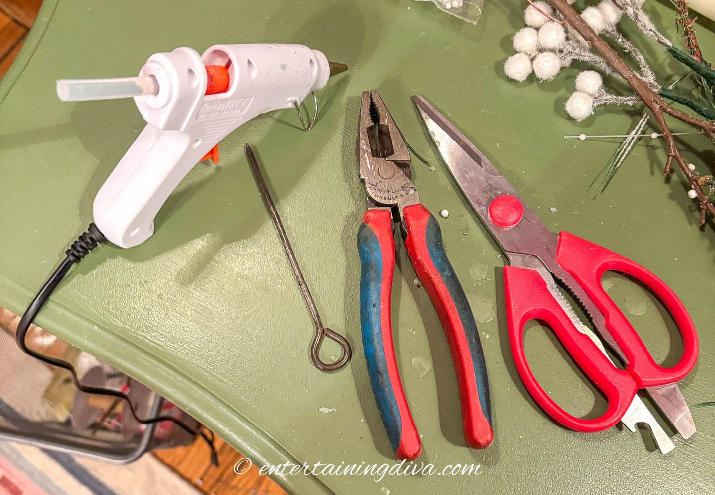 A glue gun, metal skewer, wire cutters and scissors on a table