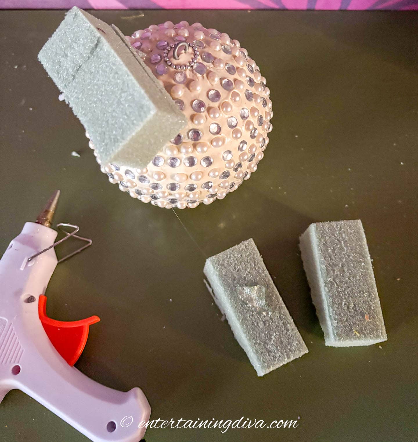 An ornament with a piece of floral foam stuck to the side of it beside a glue gun and 2 more pieces of floral foam