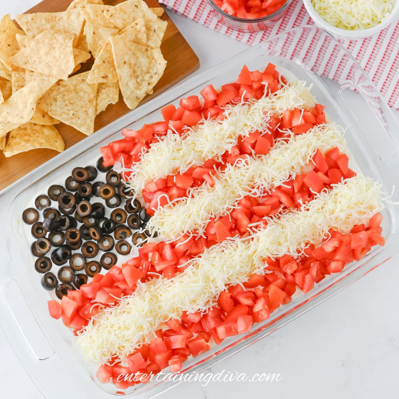 A rectangular glass dish with a taco dip decorated like the American flag using black olives, shredded cheese, and diced tomatoes, next to tortilla chips and bowls of extra toppings.