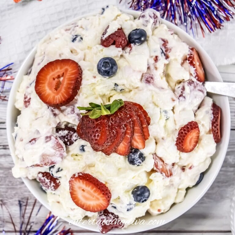 A bowl of berry cheesecake fruit salad with whipped cream, topped with sliced strawberries, blueberries, and pineapple, on a wooden surface with 4th of July decorations.