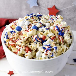 A white bowl filled with popcorn topped with red, white, and blue candy and drizzle, with star decorations in the background.