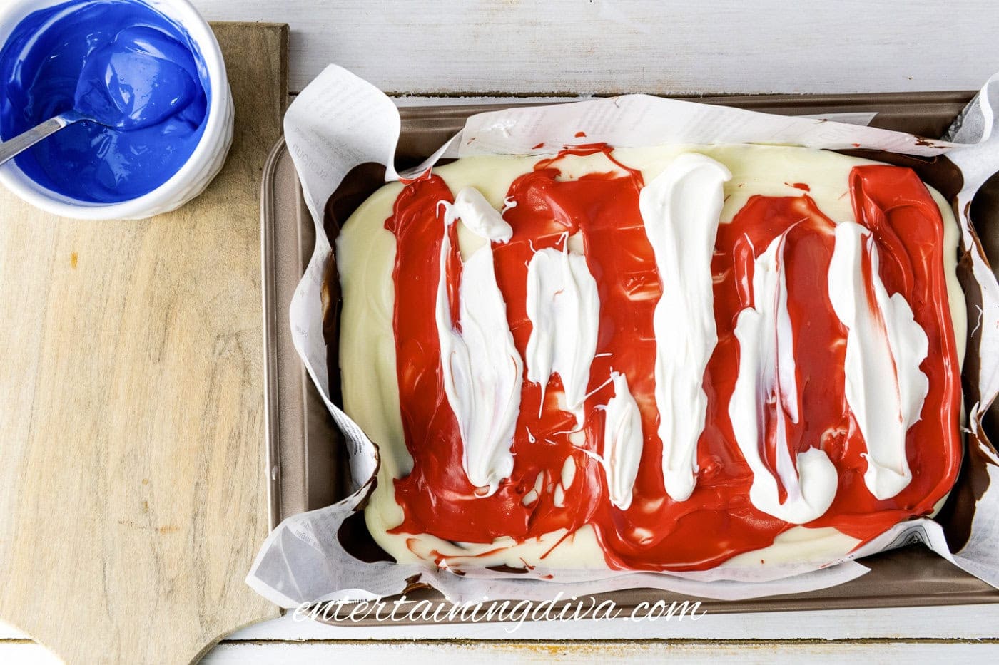 Rectangular pan with white chocolate and red candy melts swirled on top; a bowl of blue icing with a spoon sits to the side on a wooden board.