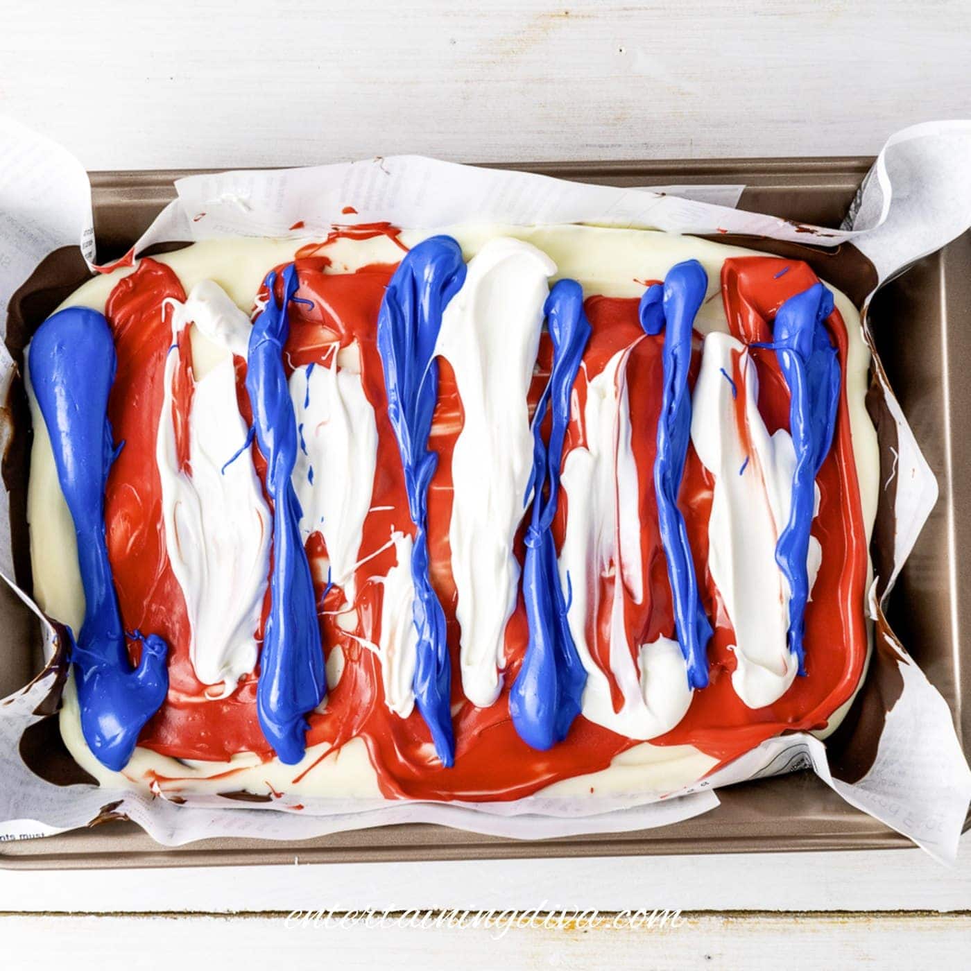 A baking pan lined with parchment paper contains melted white, red, and blue candy melts swirled together in stripes.
