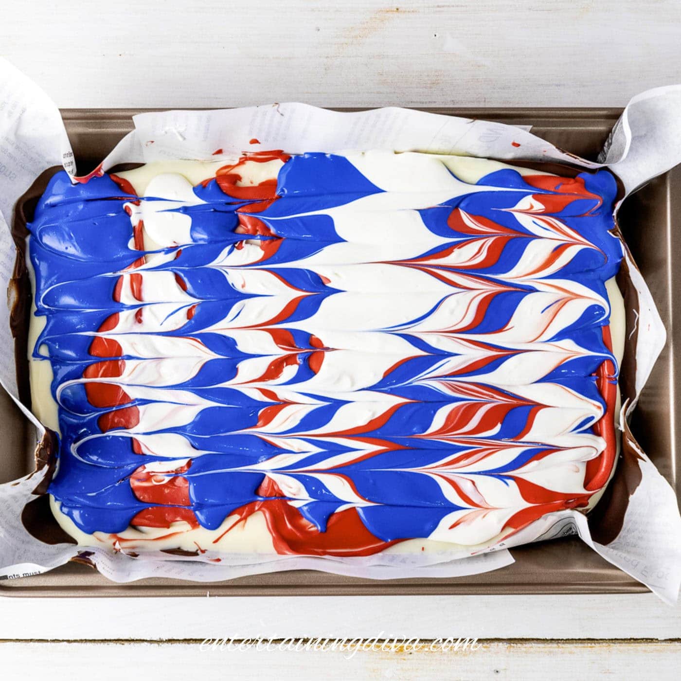 A rectangular pan lined with parchment paper holds red, white, and blue candy melts swirled in a striped pattern.