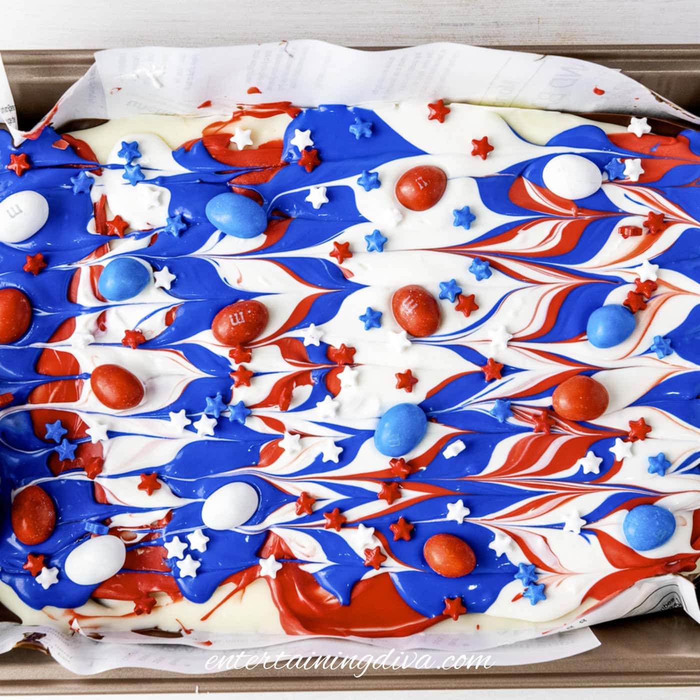 A baking tray with white chocolate bark swirled with red and blue colors, topped with red, white, and blue candies and star-shaped sprinkles.
