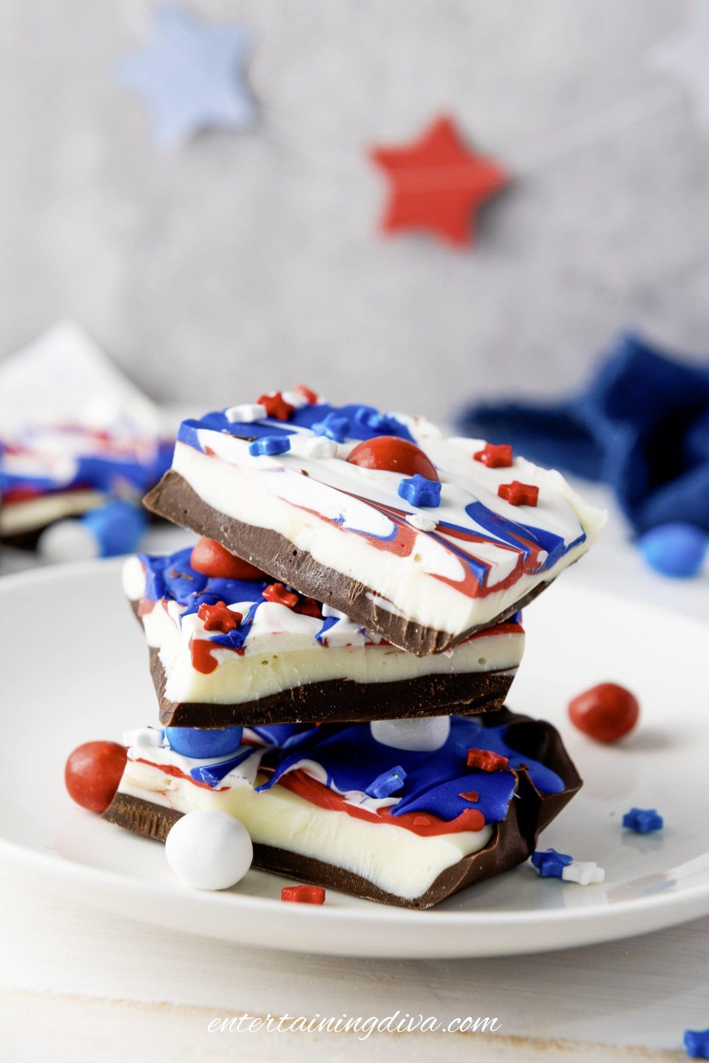 Four pieces of layered patriotic bark with red, white, and blue decorations are stacked on a white plate. Red and blue star-shaped decorations are blurred in the background.