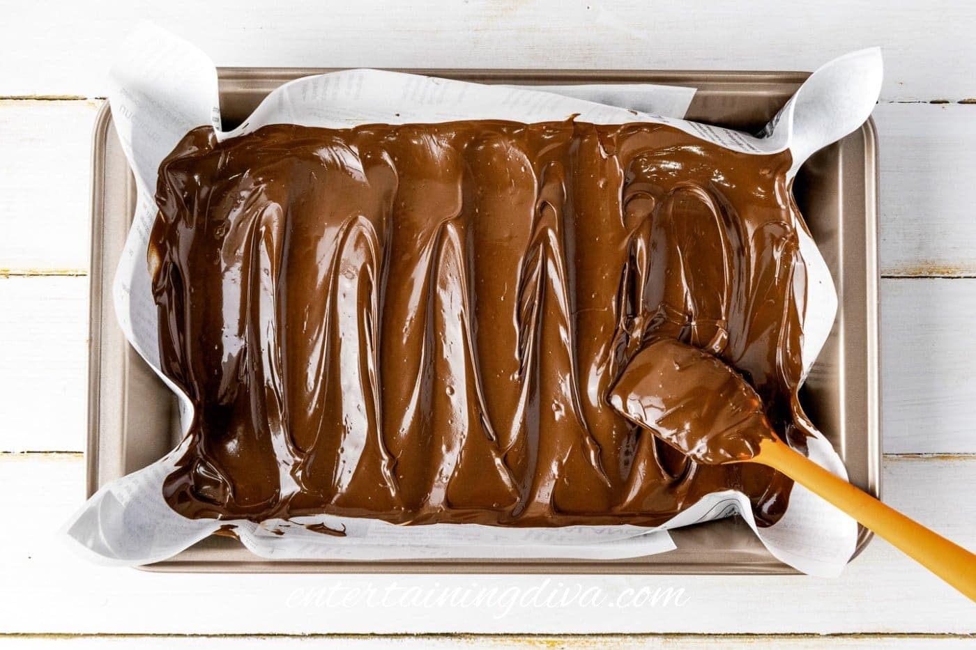 A baking tray lined with parchment paper filled with melted chocolate being spread evenly with an orange spatula.