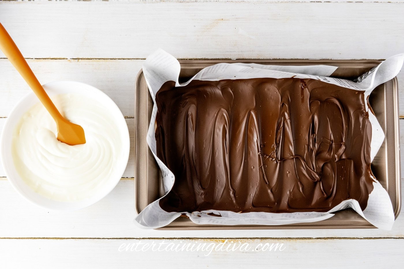 A baking tray lined with parchment paper holds a layer of chocolate. Beside it is a bowl of white frosting and an orange spatula.