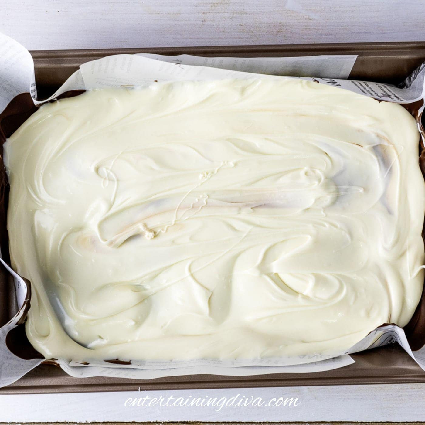 A baking pan lined with paper holds a layer of chocolate covered in a thick, even layer of swirled white chocolate.