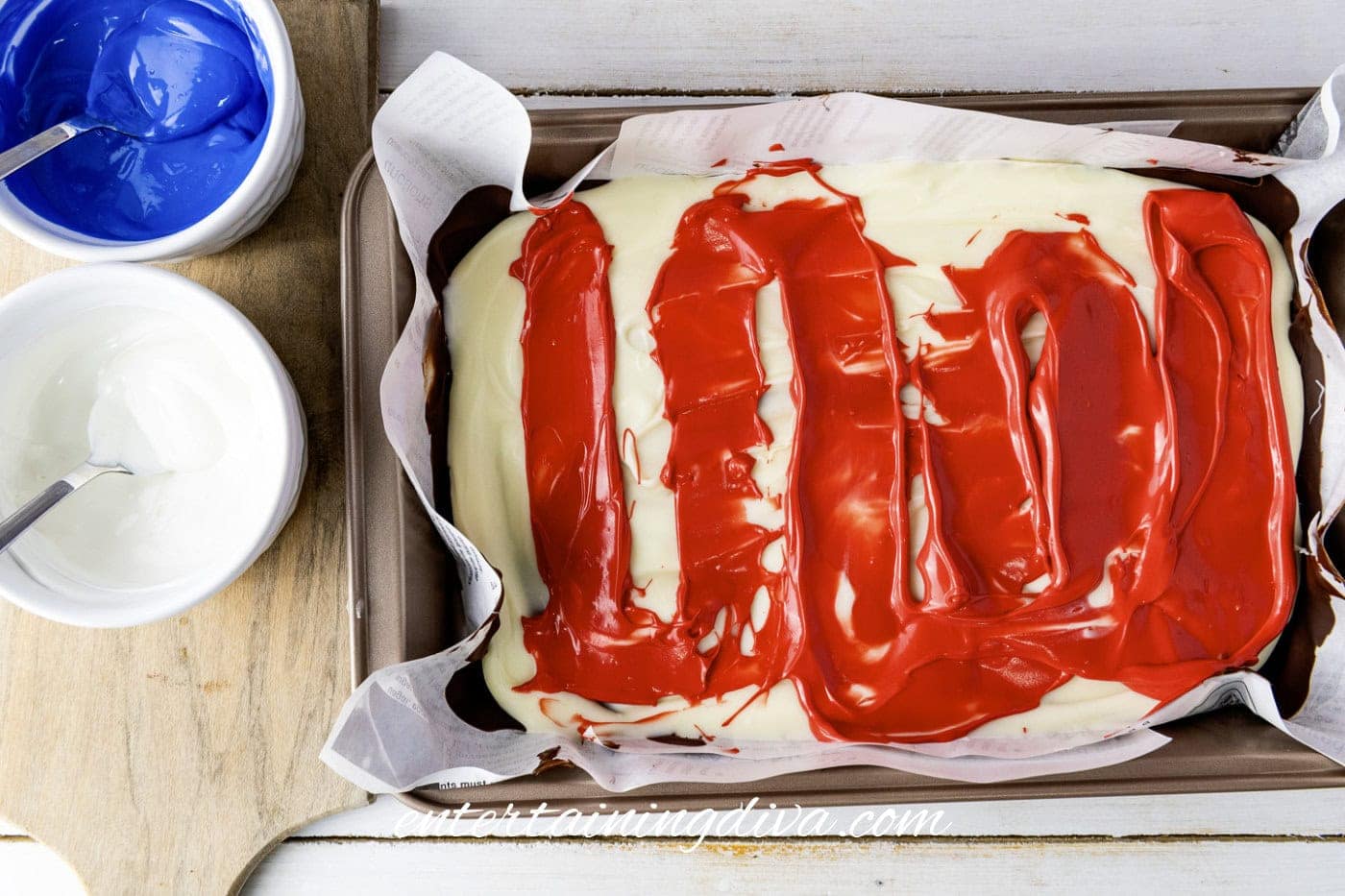 A rectangular cake in a baking pan is covered with white chocolate and red candy melts. Nearby are two bowls containing blue and white frosting with spoons.