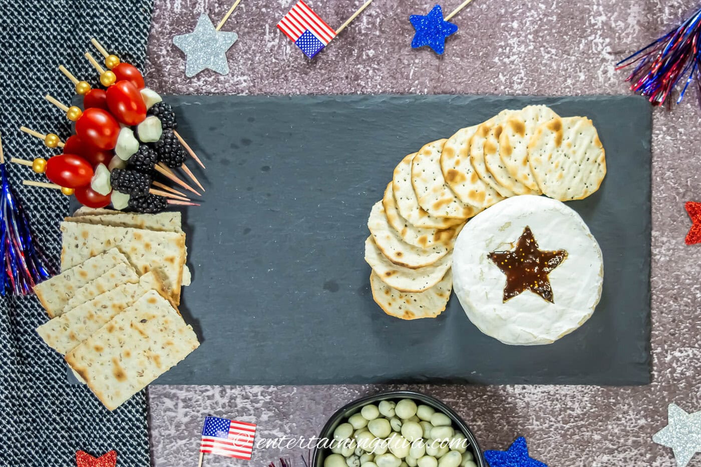 A slate board with a round of brie, round crackers, square crackers and red, white and blue skewers