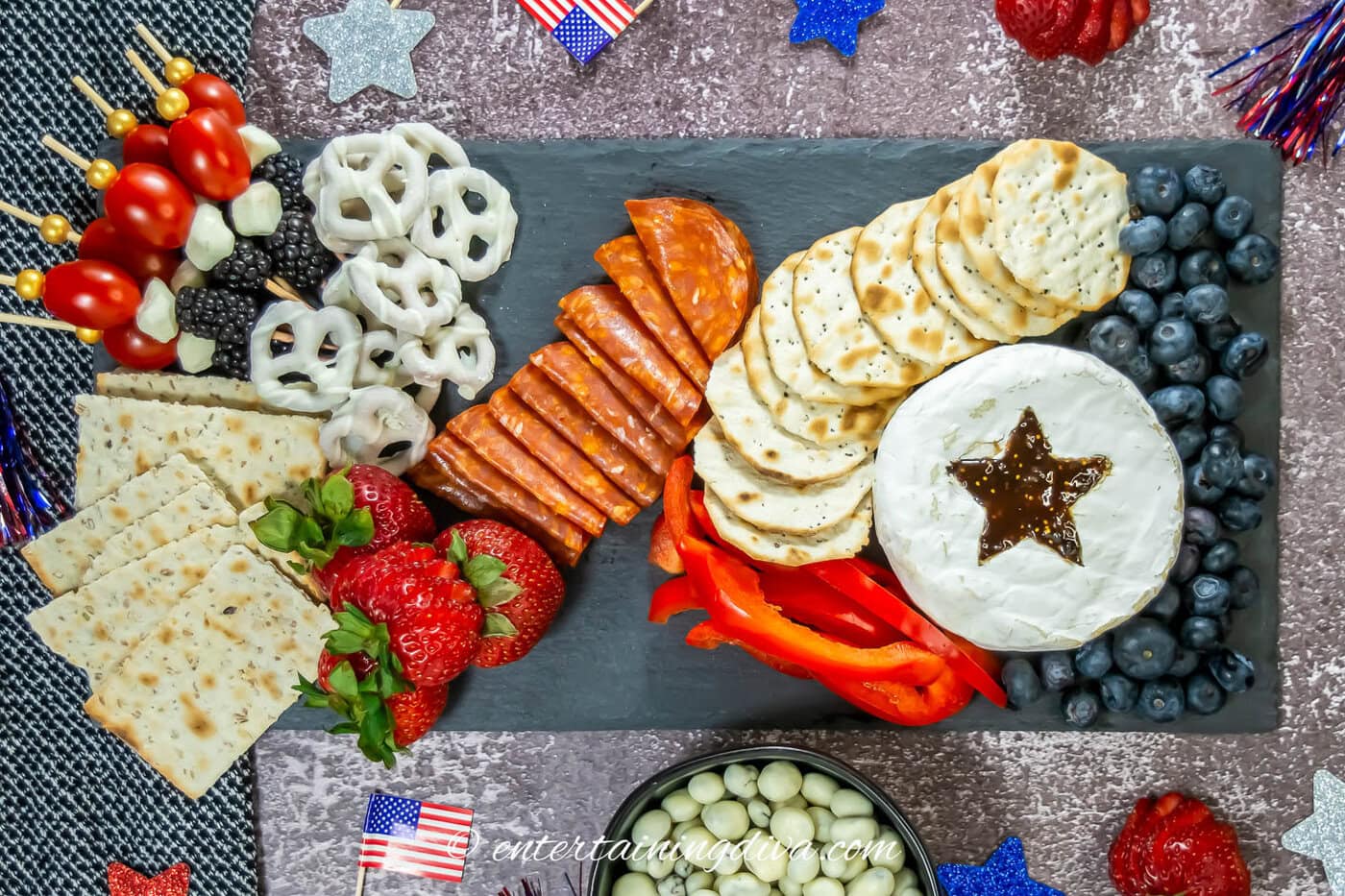 A slate board with a round of brie, round crackers, square crackers, red, white and blue skewers, red pepper slices, pepperoni slices, strawberries and white-chocolate covered pretzels