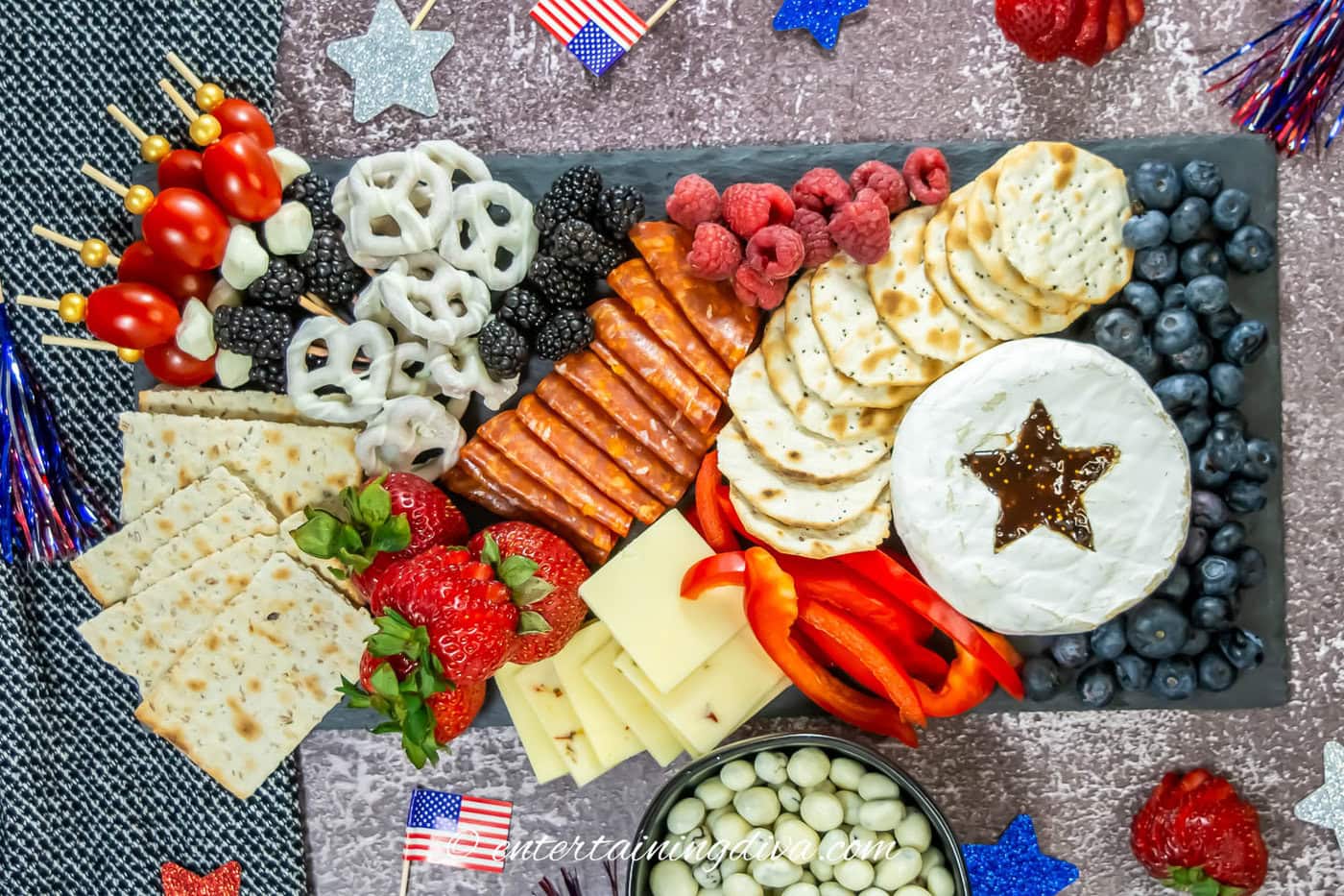A slate board filled with red, white and blue fruit, cheese and sausage.