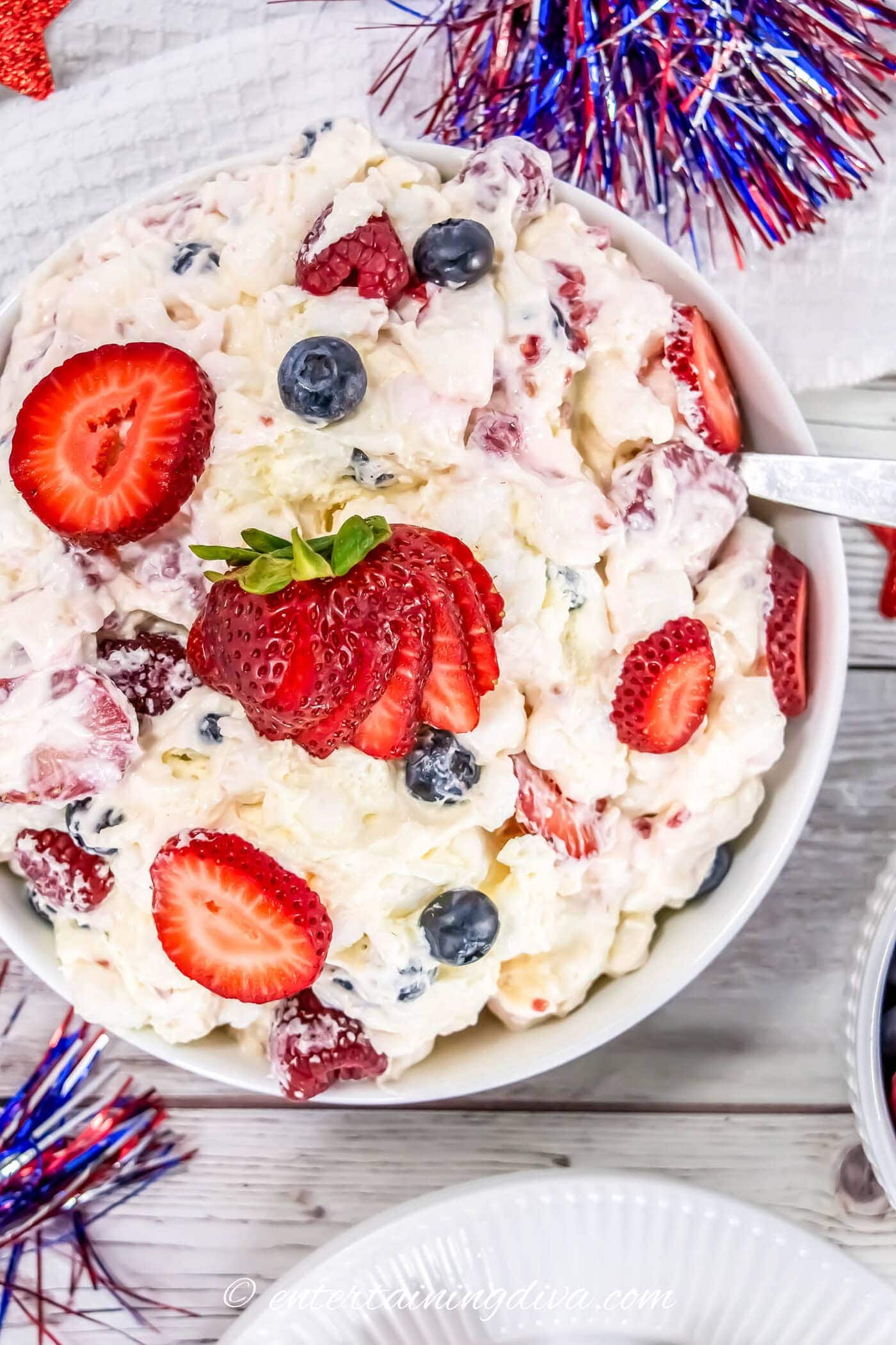 A bowl of creamy fruit salad topped with sliced strawberries and blueberries, with festive red, white, and blue decorations in the background.