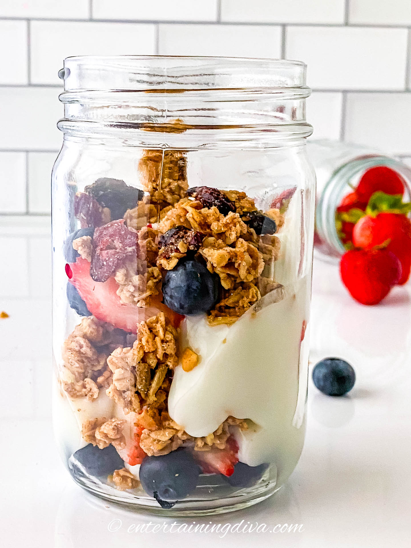 A glass jar filled with layers of yogurt, granola, strawberries, and blueberries sits on a white counter with scattered berries in the background.