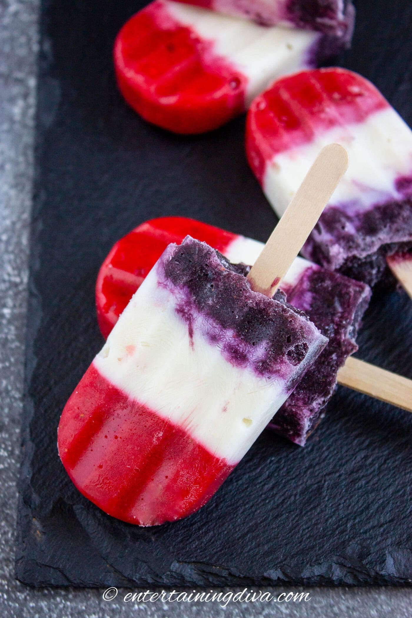 A hand holds a slice of fruit pizza topped with raspberries, blueberries, kiwi, grapes, and cream cheese frosting on a cookie crust.