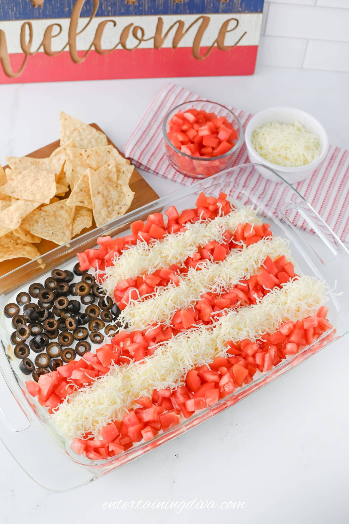 A glass dish with taco dip and an American flag design made of shredded cheese, diced tomatoes, and sliced black olives, next to tortilla chips and small bowls of extra toppings.