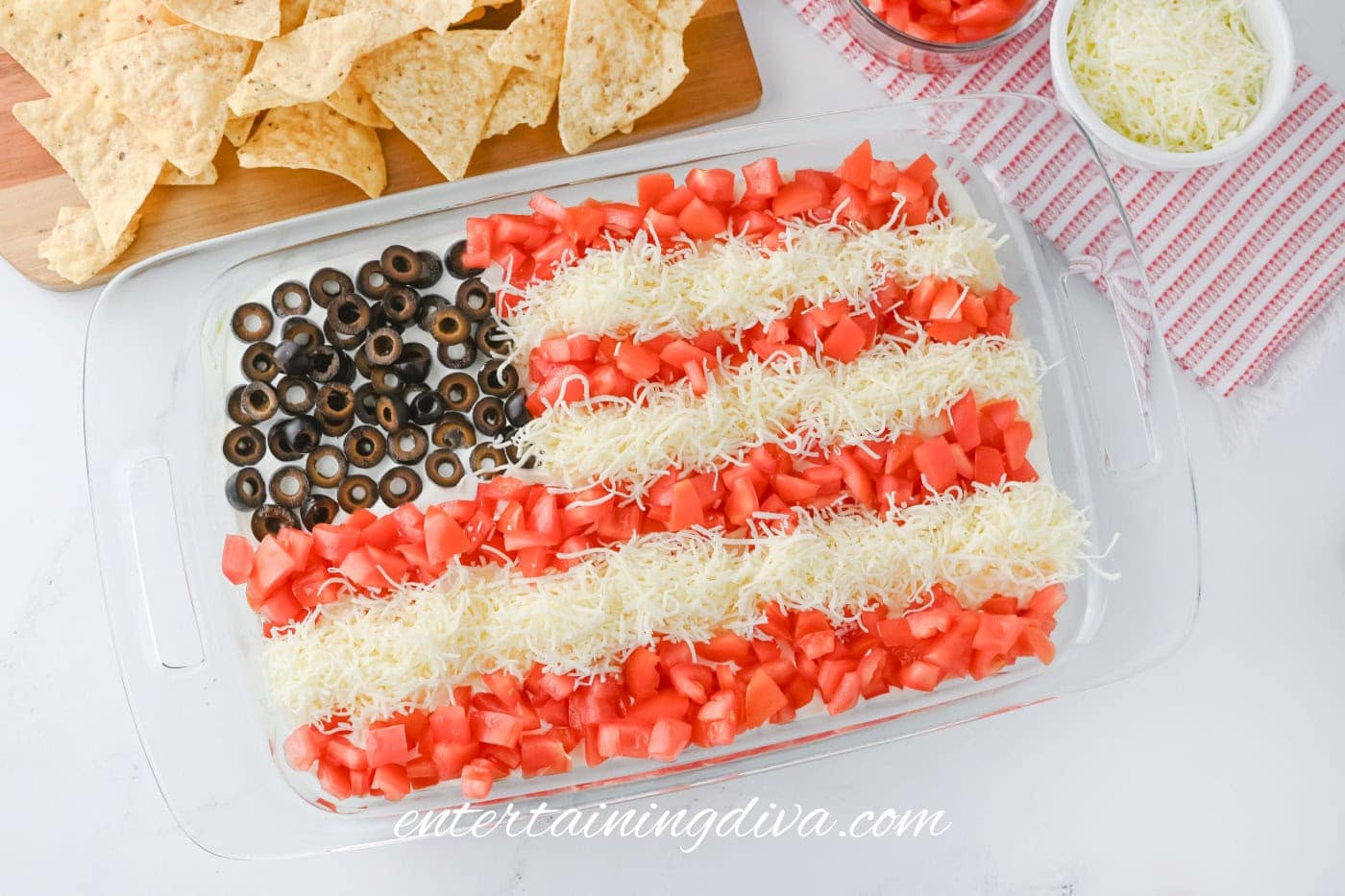 A glass dish with a layered taco dip arranged as an American flag using olives, tomatoes, and shredded cheese. Tortilla chips and bowls with cheese and tomatoes are nearby.