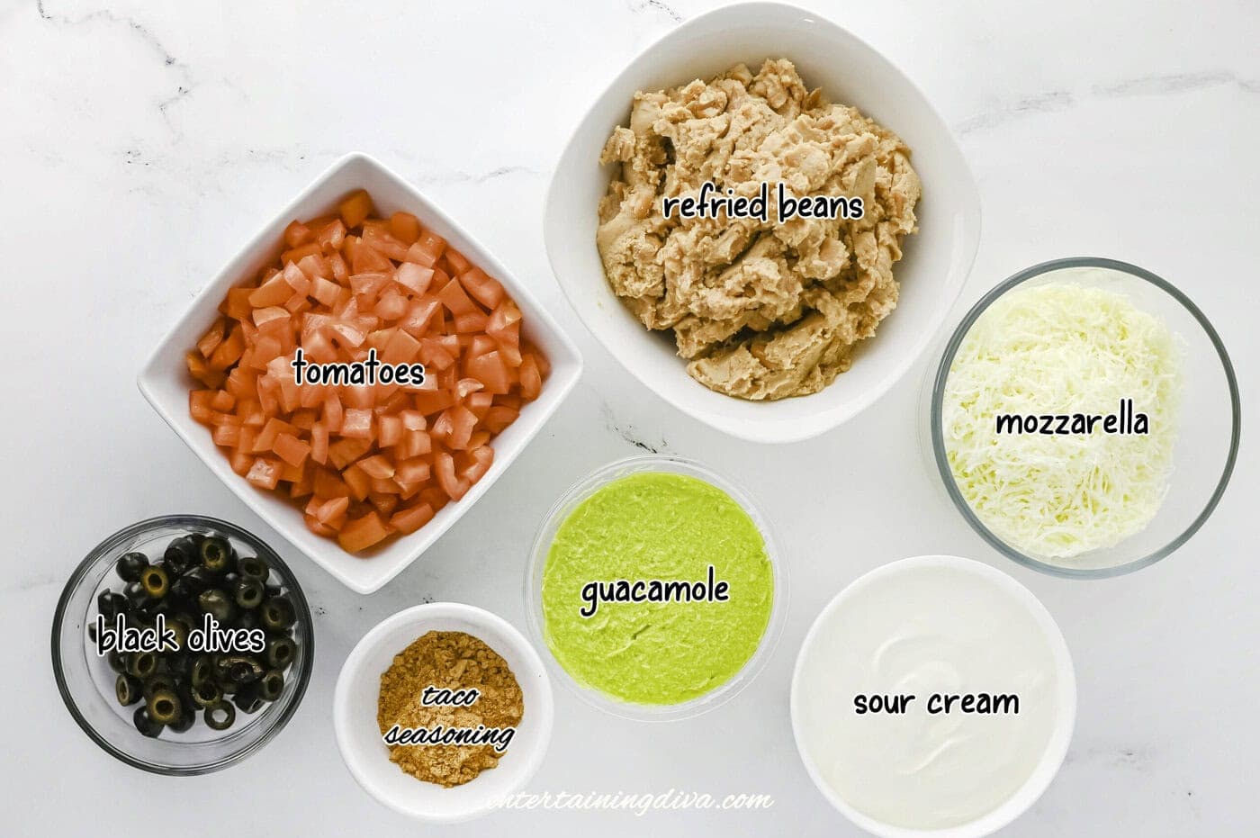 Seven bowls containing refried beans, diced tomatoes, shredded mozzarella, sliced black olives, taco seasoning, guacamole, and sour cream are arranged on a white surface.