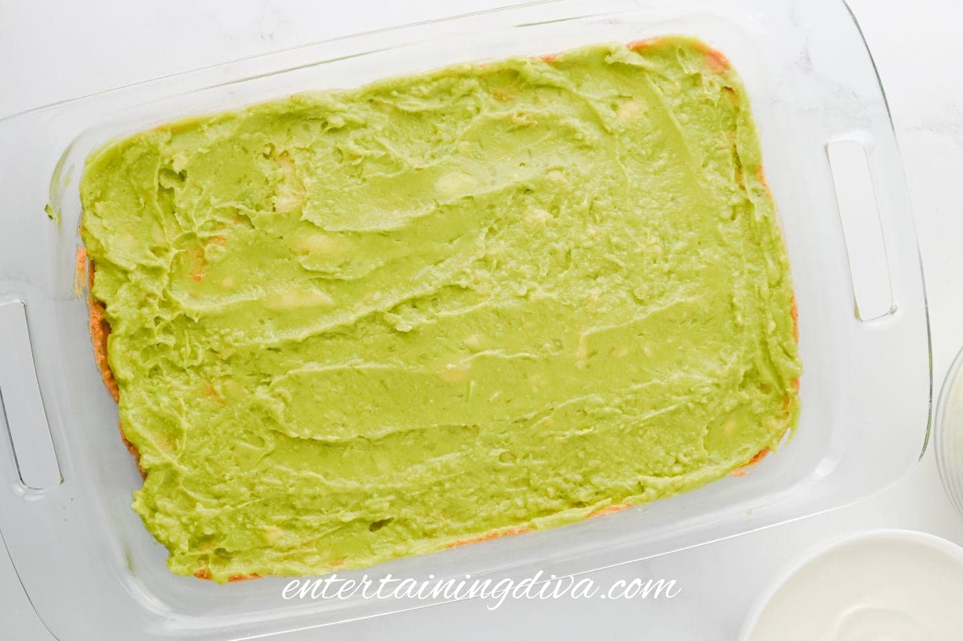 A glass baking dish filled with a smooth layer of guacamole spread evenly across the surface of the refried beans.