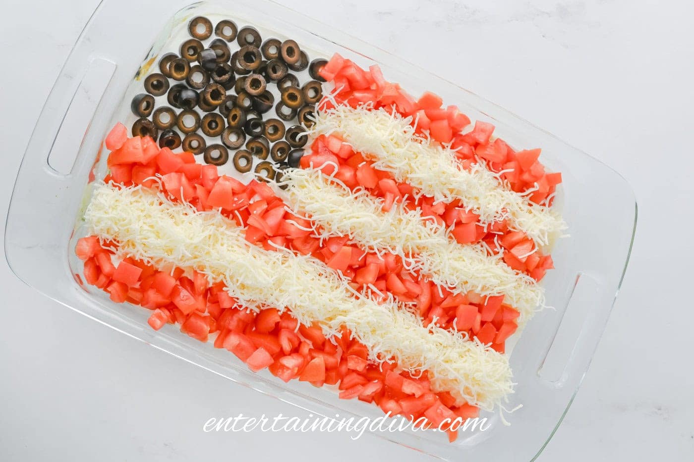A glass dish contains a layered taco dip topped with chopped tomatoes, shredded cheese in stripes, and sliced black olives in the corner, arranged to resemble the American flag.