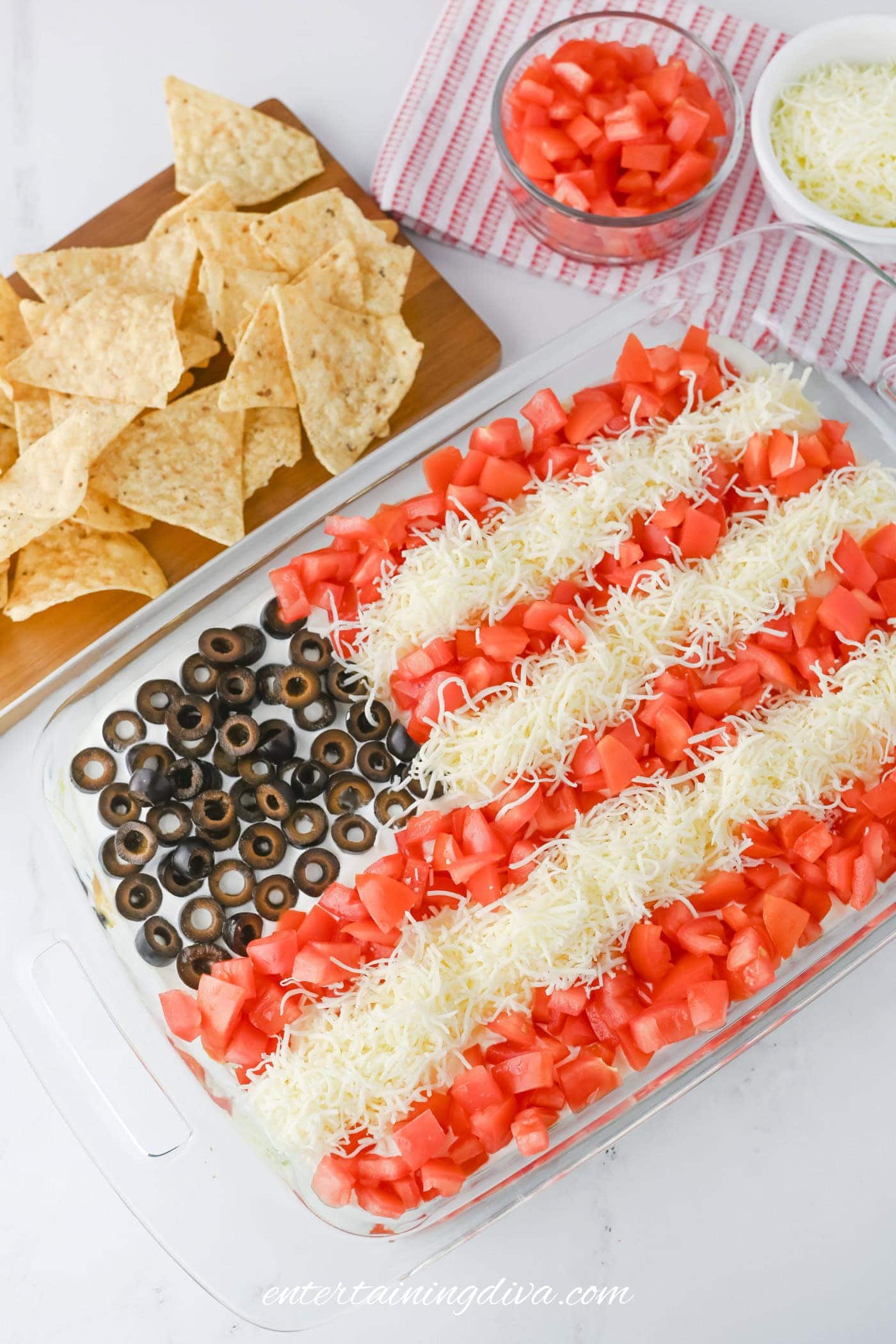 A rectangular dip in a glass dish is decorated with black olives, shredded cheese, and diced tomatoes to resemble the American flag, accompanied by tortilla chips on the side.