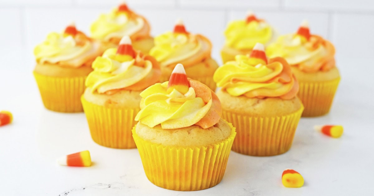 Seven cupcakes with yellow and orange swirled frosting, each topped with a piece of candy corn, are arranged on a white surface with a few loose candy corn pieces nearby.