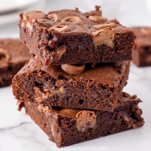 Three chocolate brownies stacked on a white surface, showing a rich, moist texture with visible chocolate chunks.