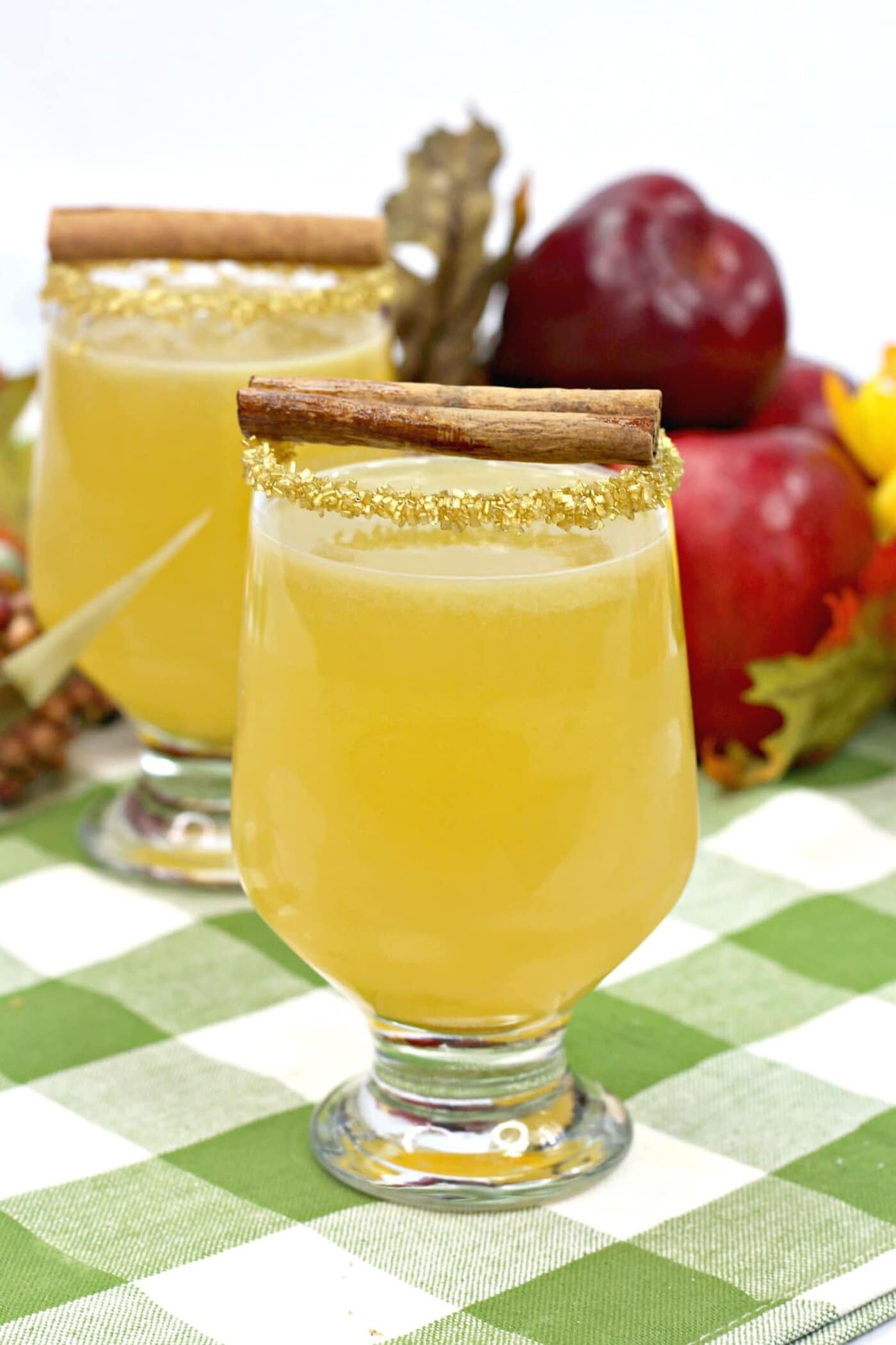 Two glasses of apple cider with cinnamon sticks on a checkered tablecloth.