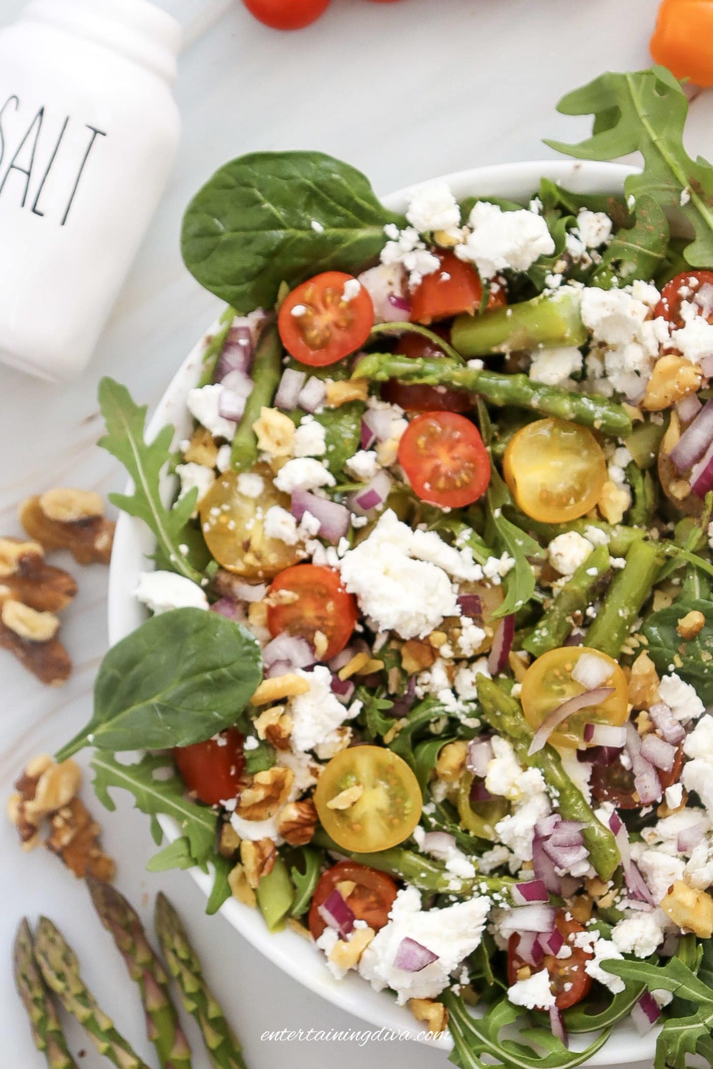 A bowl of fresh salad with cherry tomatoes, spinach, arugula, red onion, walnuts, feta cheese, and asparagus, with a salt shaker nearby.