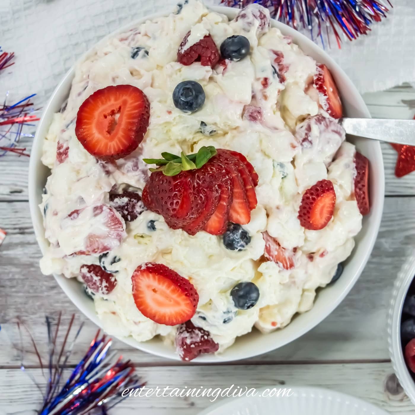 A bowl of fruit salad with whipped cream, strawberries, and blueberries on a light wooden table with red, white, and blue festive decorations.