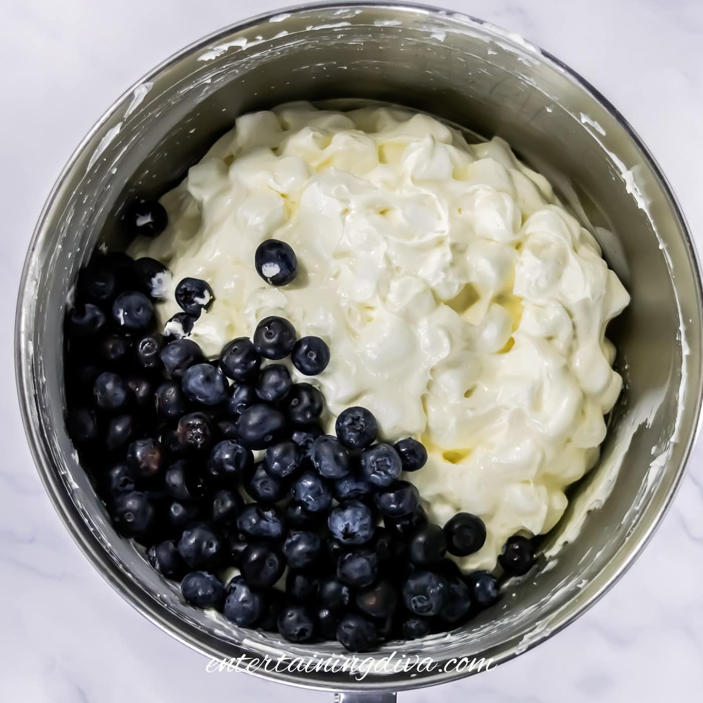 A mixing bowl containing a cheesecake mixture with fresh blueberries on one side, ready to be stirred together.