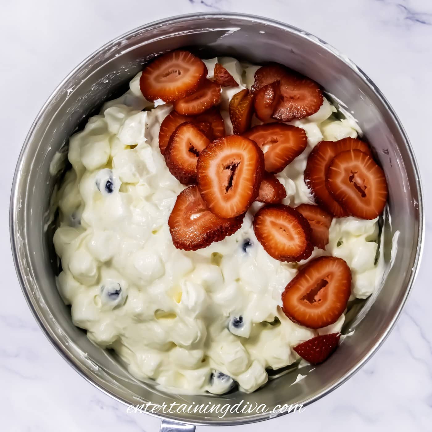 A mixing bowl filled with cheesecake fruit salad containing marshmallows, blueberries, and topped with sliced strawberries.