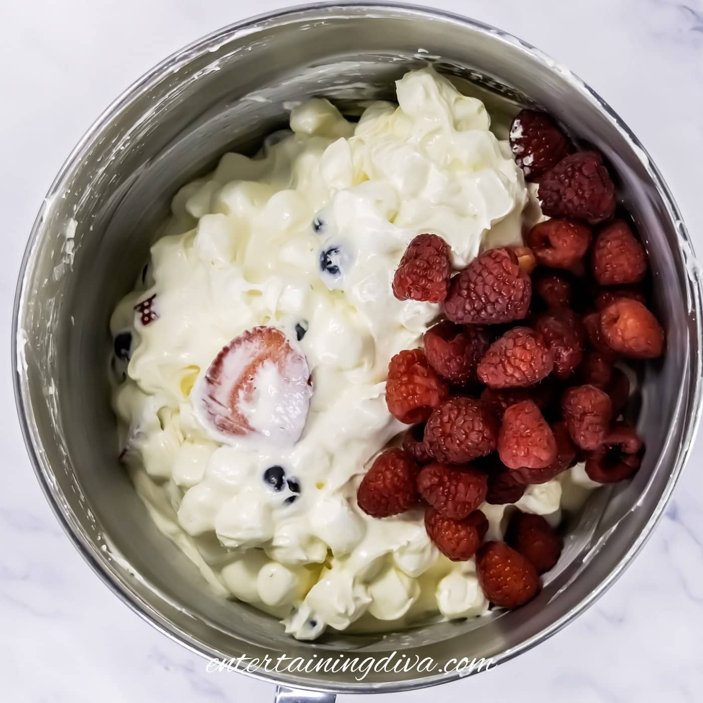 A mixing bowl containing cheesecake fruit salad with marshmallows, blueberries, sliced strawberries, and fresh raspberries.