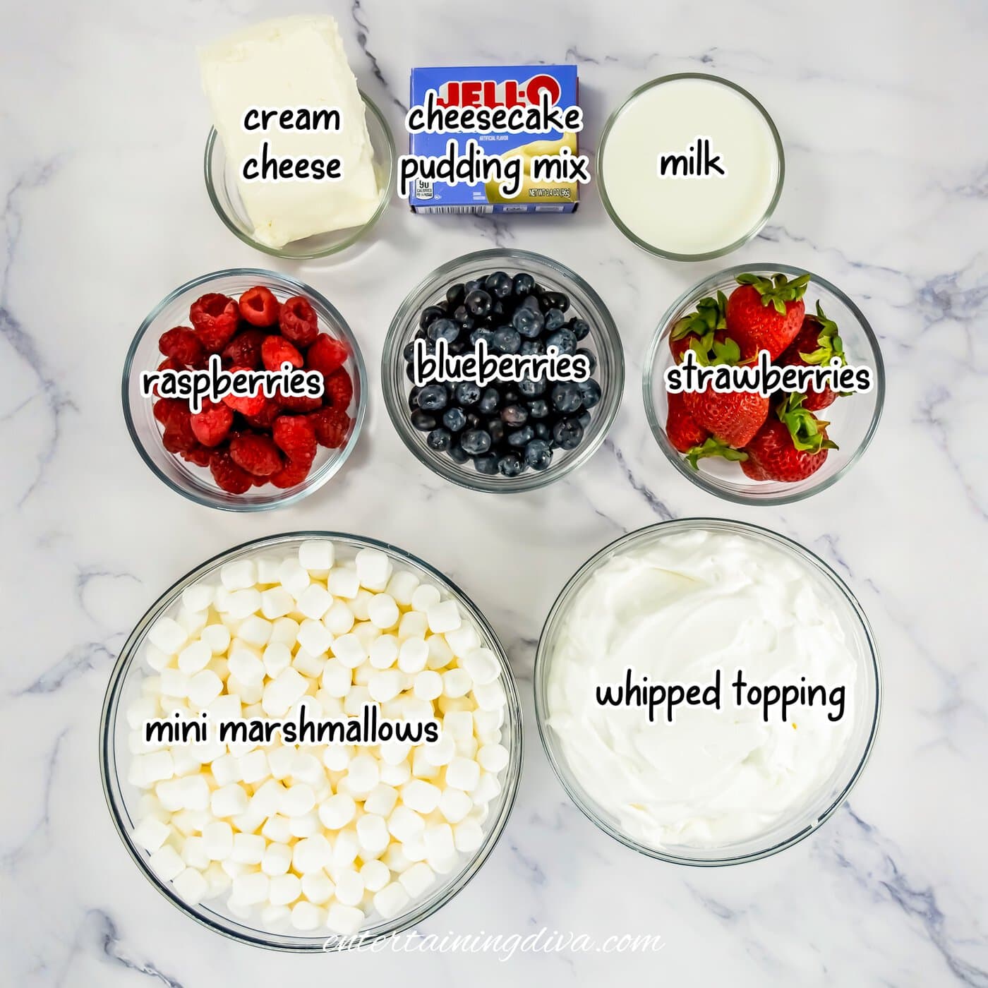 Top-down view of bowls containing cream cheese, cheesecake pudding mix, milk, raspberries, blueberries, strawberries, mini marshmallows, and whipped topping on a marble surface.